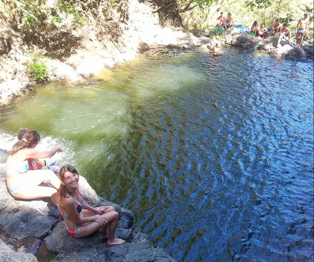 A clear jungle swimming hole below a waterfall, with swimmers resting on the rocks